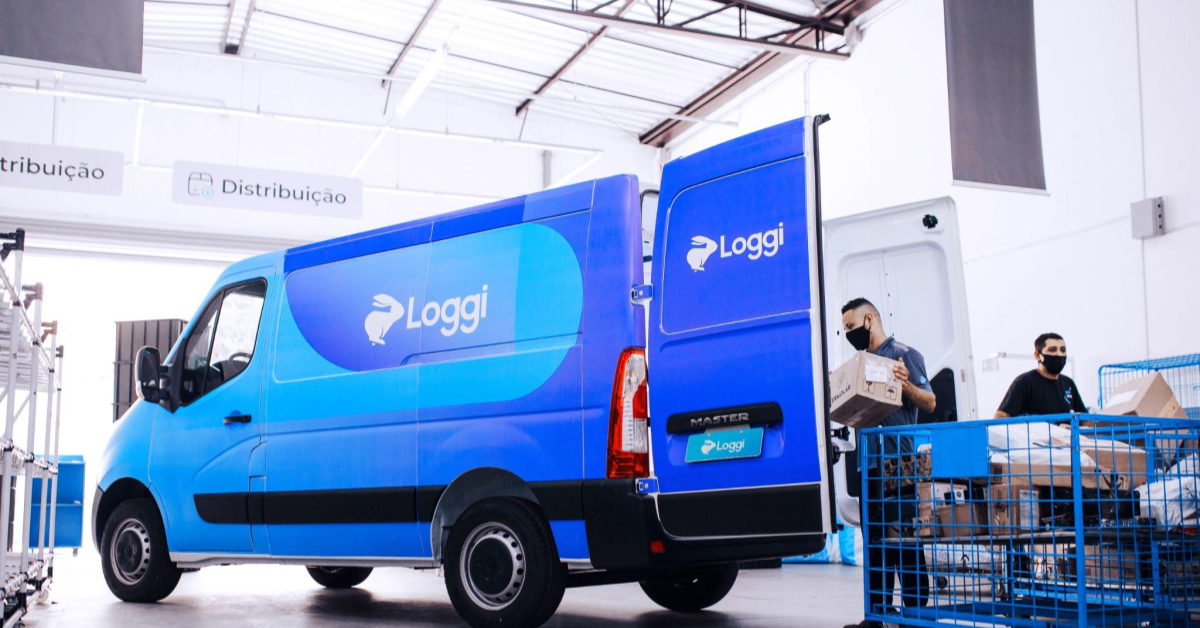 A imagem mostra uma das vans da Loggi sendo carregada com pacotes por um homem de camisa preta. A van está localizada em um dos crossdockings da Loggi.
