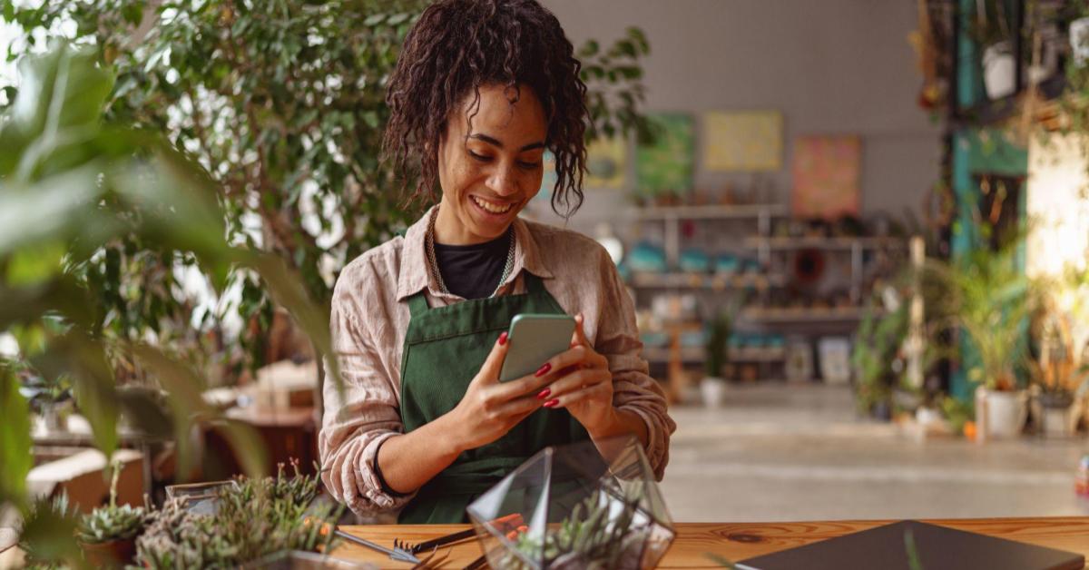 Na imagem, uma mulher negra, empreendedora, está em uma loja de flores. O foco da imagem está na mulher que usa seu celular para tirar a foto de uma de suas plantas. Um exemplo de como fotografar produtos para o e-commerce.