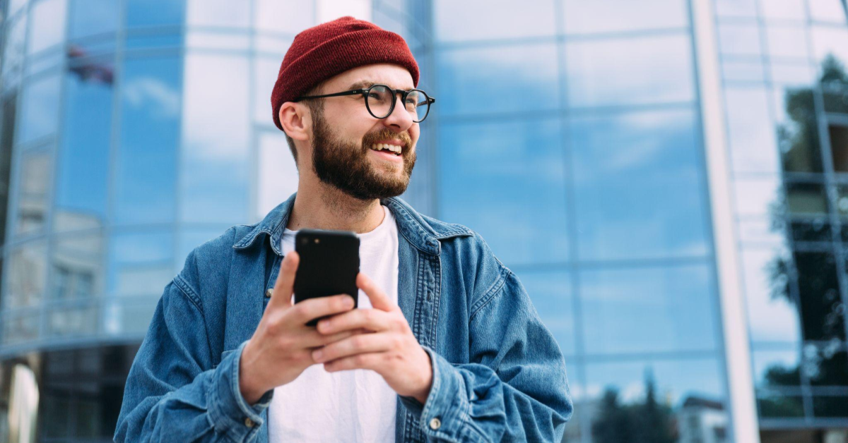 Um homem branco, usando óculos e vestindo gorro vermelho, camiseta branca e camisa jeans azul, está segurando um celular enquanto olha na direção direita da imagem e sorri. O celular aqui acaba sendo um dos dispositivos usados para o social commerce