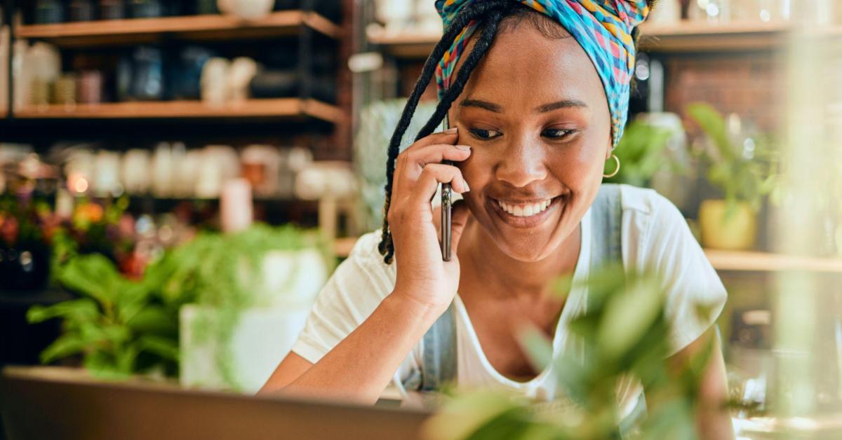 Uma mulher negra usa dreads e faixa colorida no cabelo, camiseta branca e um macacão jeans. Na imagem, ela sorri enquanto fala no celular. Ela está rodeada por plantas, possivelmente em uma floricultura. 