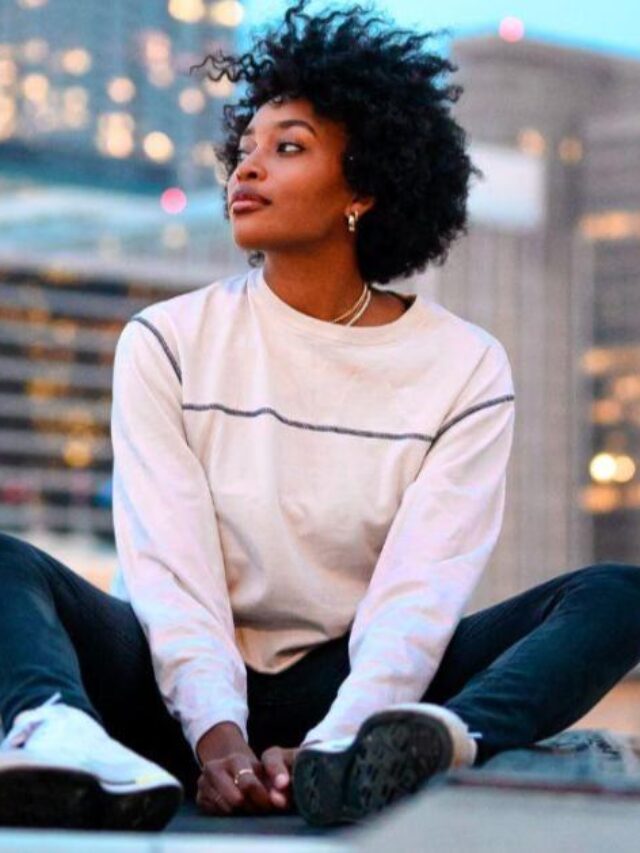 Imagem de uma mulher preta sentada na sacada de um prédio. A mulher tem cabelos encaracolados, veste uma blusa branca de manga longa, calça jeans escura e tênis brancos. Ao fundo, é possível ver as luzes de diversos prédios. A imagem foi feita por Jeffery Erhunse no Unsplash, e faz alusão aos influenciadores digitais.
