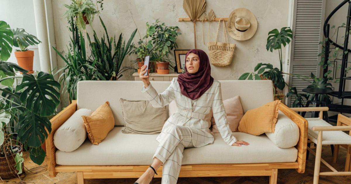 A imagem mostra uma mulher, que usa terno branco e um hijab vinho. Ela está sentada em um sofá branco, enquanto tira fotos com seu celular. Uma alusão ao momento em que pessoas que querem vender no instagram se programam para fotografar seus produtos.
