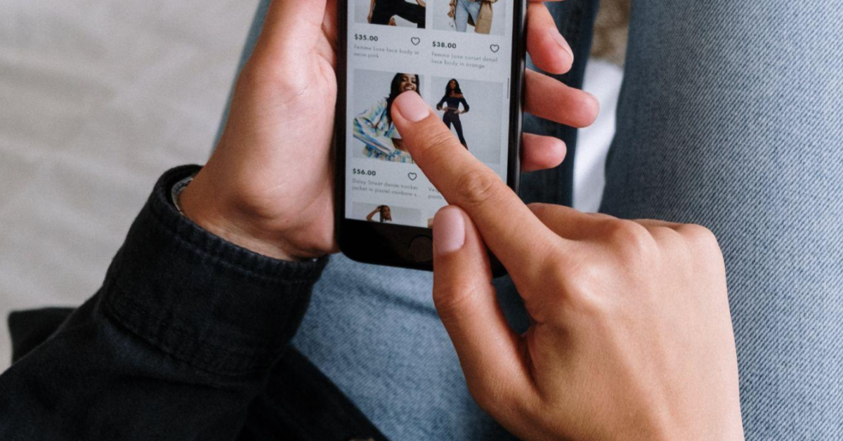 A imagem foca na mão de uma pessoa, de pele negra, mexendo no celular, mais especificamente, em uma seção de roupas de um e-commerce, uma alusão aos clientes que buscam pelas promoções das datas duplas da shopee