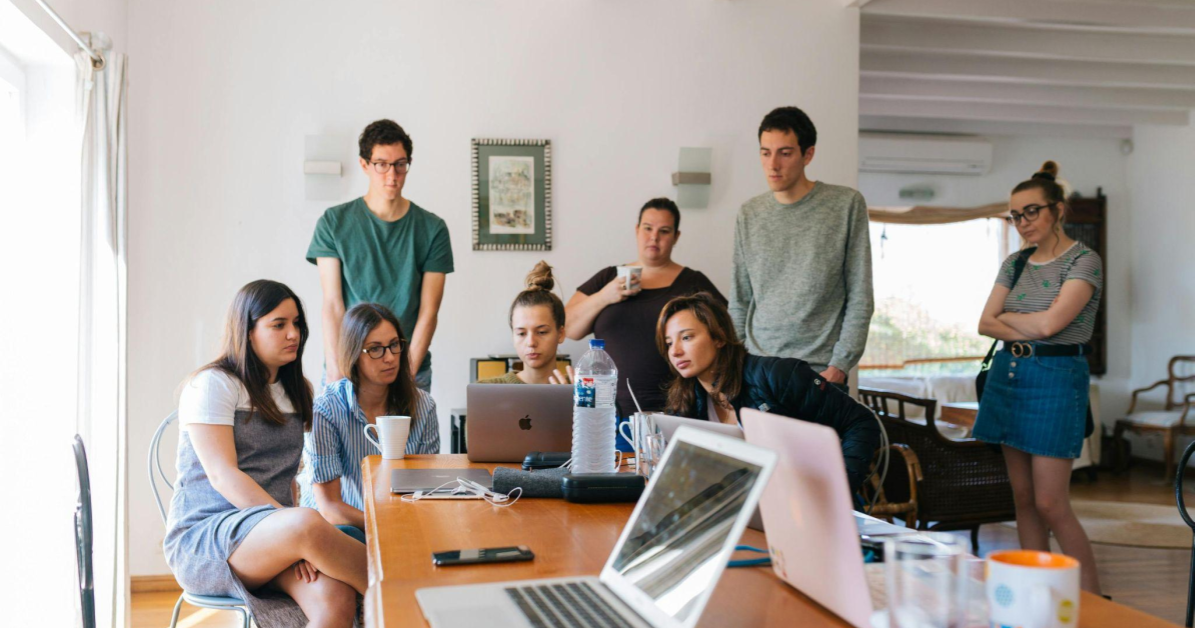 A imagem mostra um grupo de pessoas, homens e mulheres, reunidos em um escritório. As pessoas estão sentadas em torno de uma mesa de trabalho, com computadores ligados, conversando sobre estratégias de aprimoração do atendimento ao cliente.