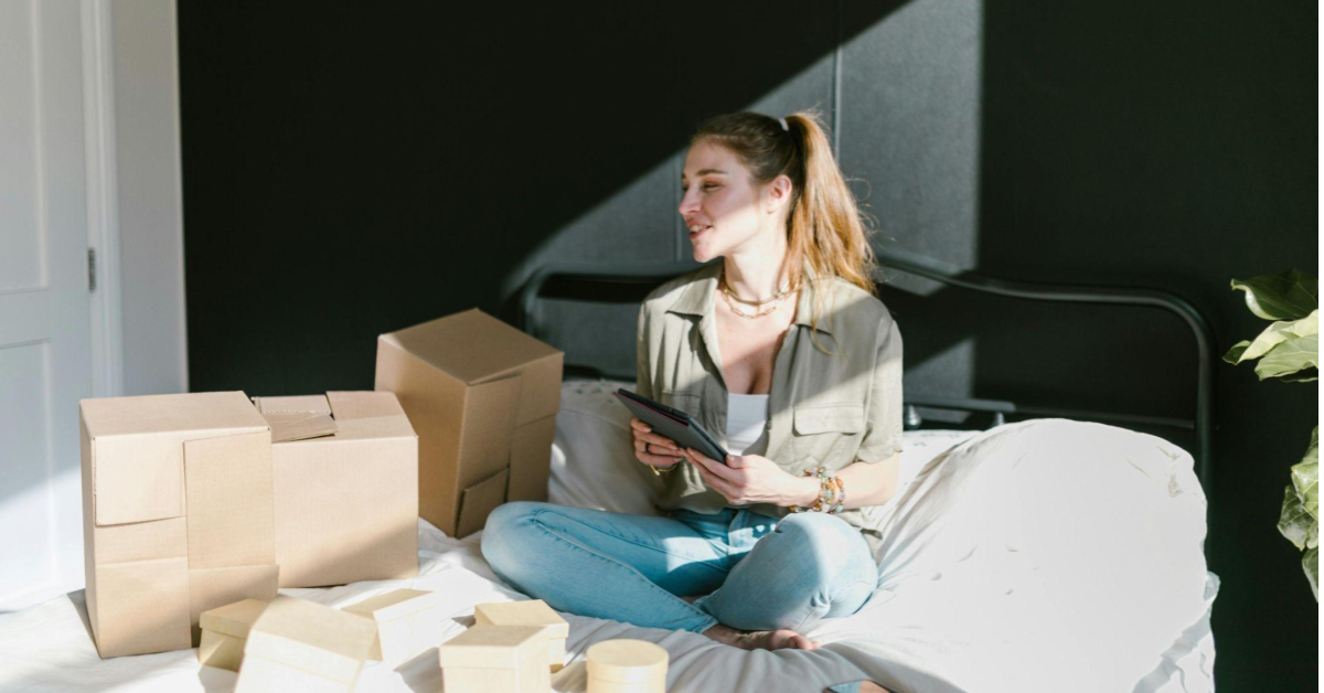 A imagem mostra uma mulher branca, loira, de cabelos amarrados. Ela está sentada em uma cama, mexendo em pacotes. Uma alusão a um momento em que uma pessoa empreendedora se organiza para vender na Amazon.