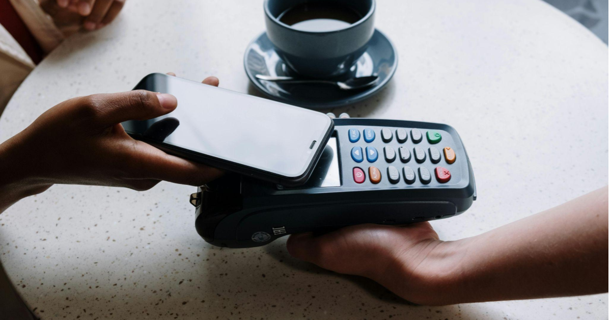 a imagem mostra a interação de duas mãos, sendo que uma segura um celular e outra segura uma máquina de pagamentos, uma alusão ao uso de gateway de pagamento no e-commerce.