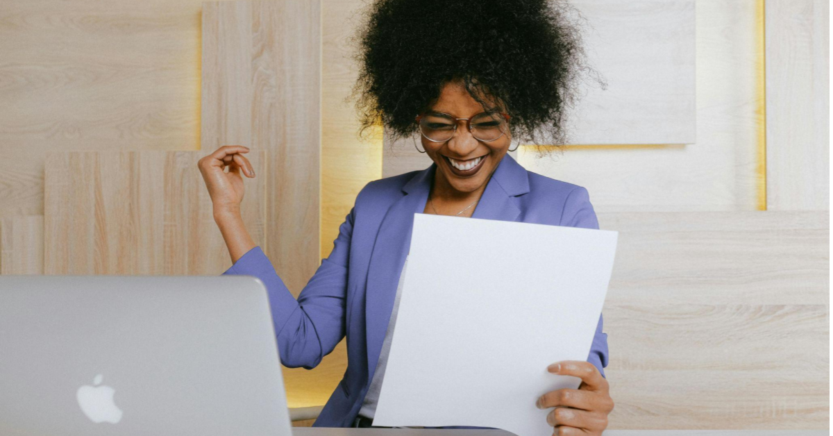 A imagem mostra uma mulher negra, de cabelos encaracolados, vestindo uma camisa social roxa clara. Ela está sentada numa cadeira, de frente para uma mesa. Na imagem, ela segura um papel (RPA) e sorri como se estivesse comemorando.