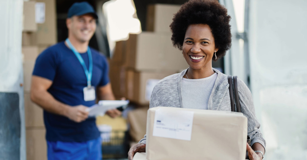 Cliente sorridente recebendo encomenda de uma das melhores transportadoras do Brasil, com entregador ao fundo e van de entregas carregada.