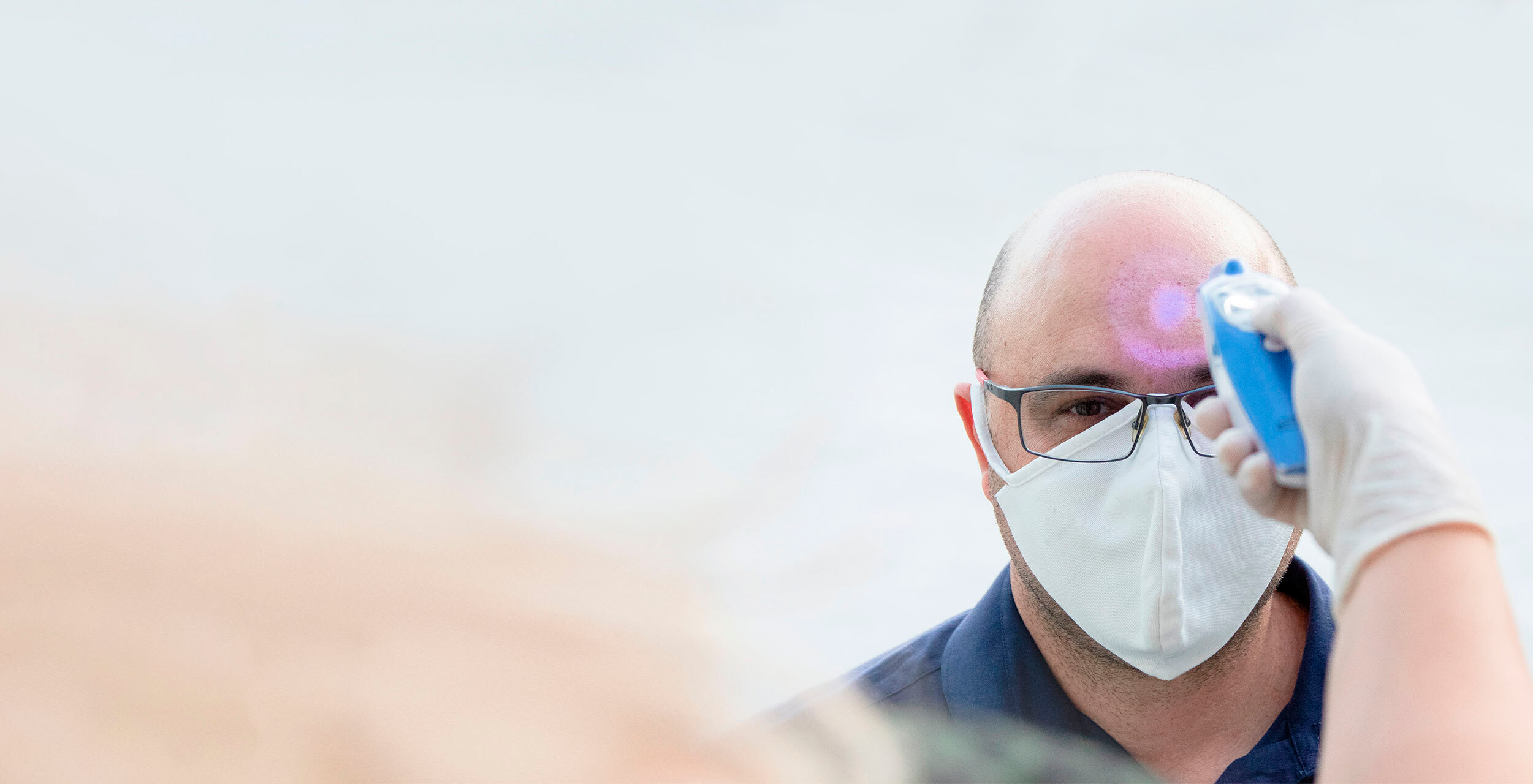 Homem branco de óculos e de máscara tendo a sua temperatura medida